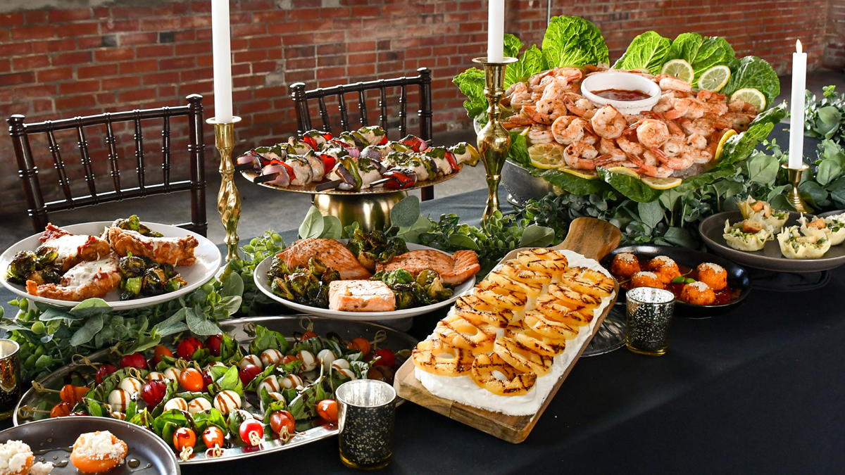 Catering spread with shrimp cocktail, chicken skewers, grilled pineapple, burrata cheese, salmon, and arancini